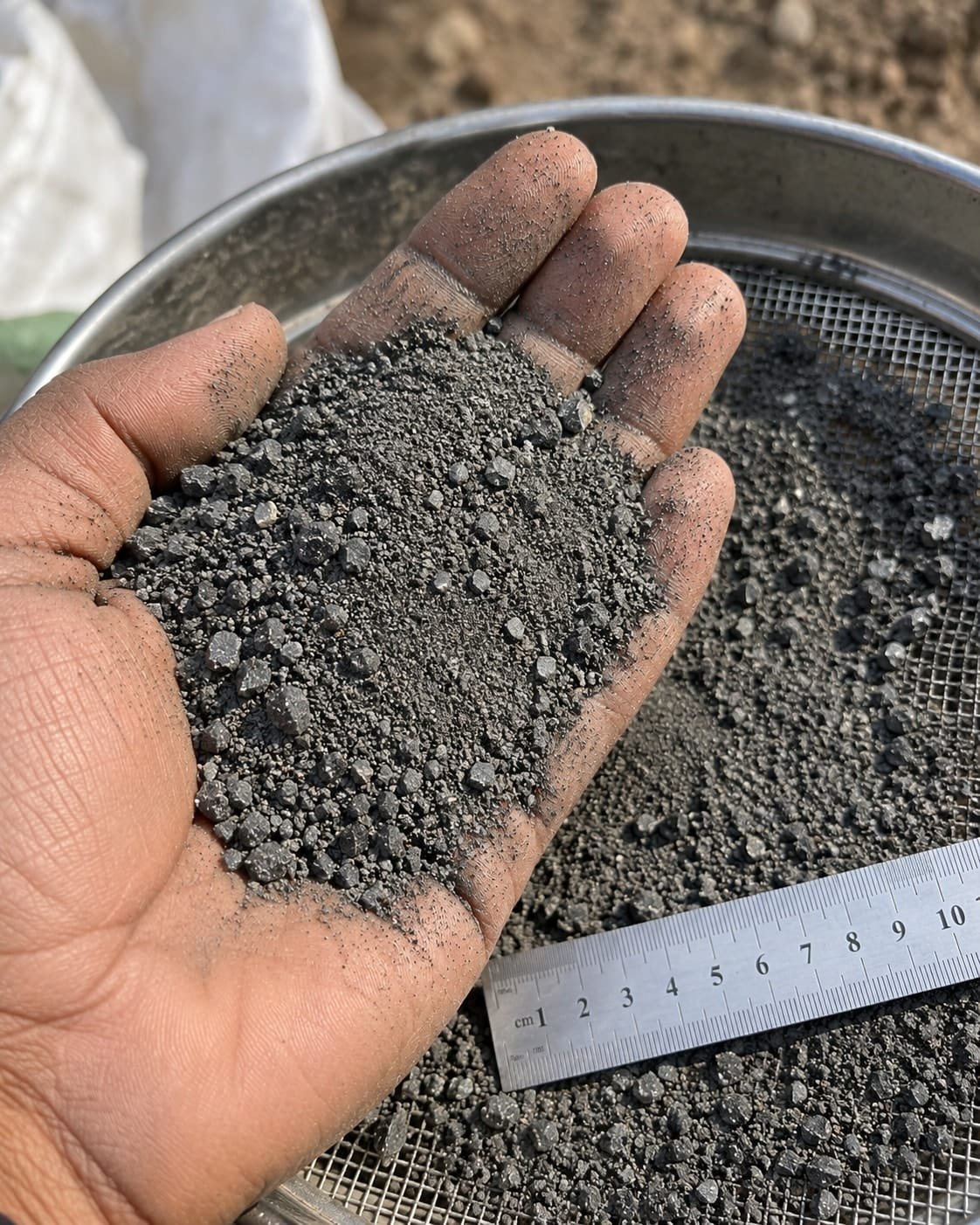 An open palm holding crushed dark-grey basalt over a steel sieve: angular grains from fine dust to pieces several millimetres across, dust on the fingers, and a centimetre ruler laid across the sieve for scale.