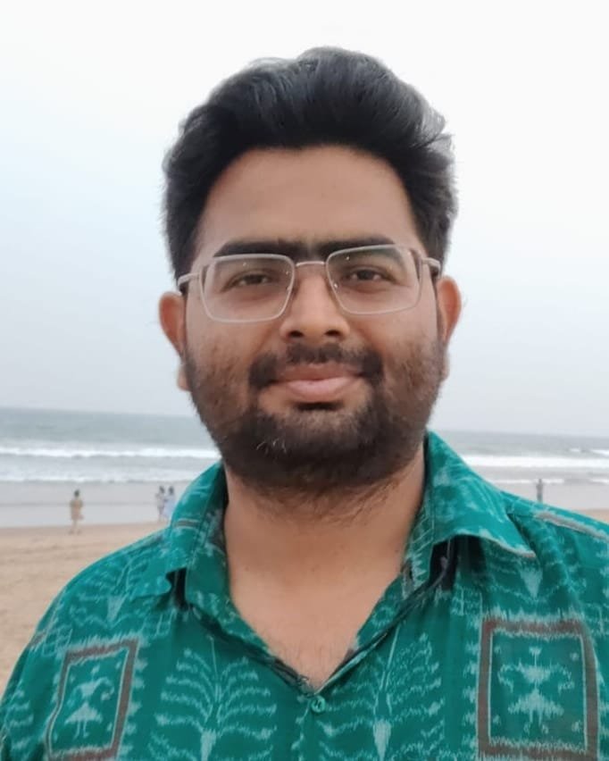 Biswajit Panda on a beach under an overcast sky, in glasses and a green ikat-print shirt, the surf and wet sand behind him.