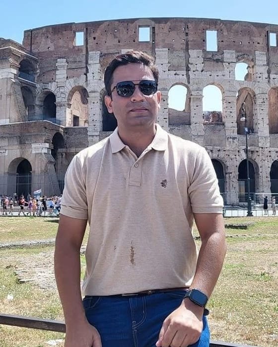 Dr. Imran Khan in sunlight in front of the Colosseum in Rome, in sunglasses and a beige polo shirt, leaning against a railing.