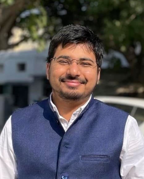 Dr. Juzer Shaikh outdoors in daylight, smiling in rimless glasses and a navy waistcoat over a white shirt, trees and a white building blurred behind him.