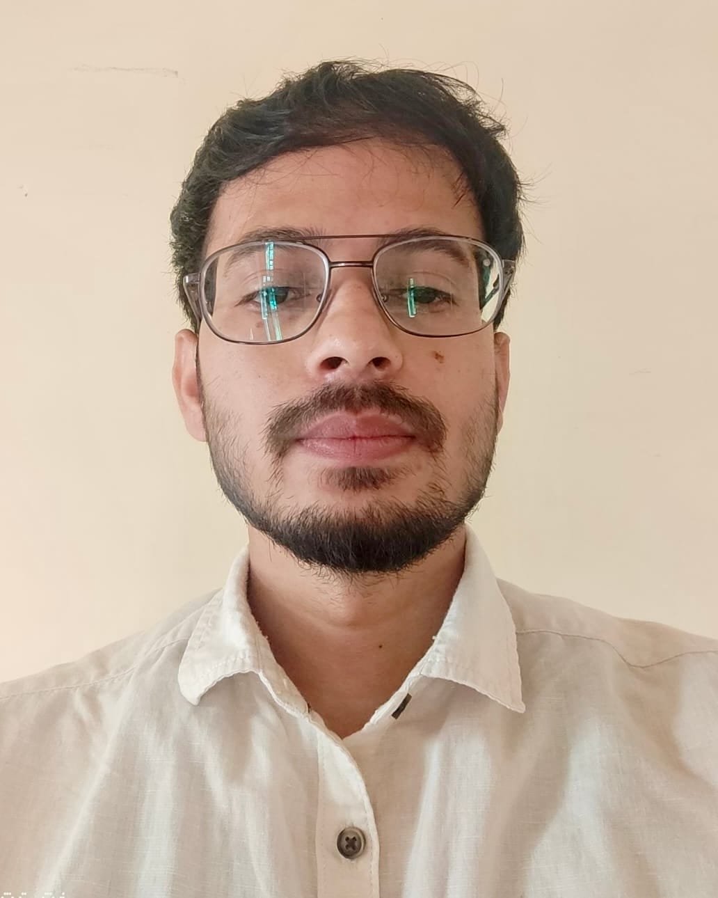 Nihal-ud-din indoors against a plain cream wall, in wire-framed glasses and an open-collared off-white linen shirt, looking straight at the camera.
